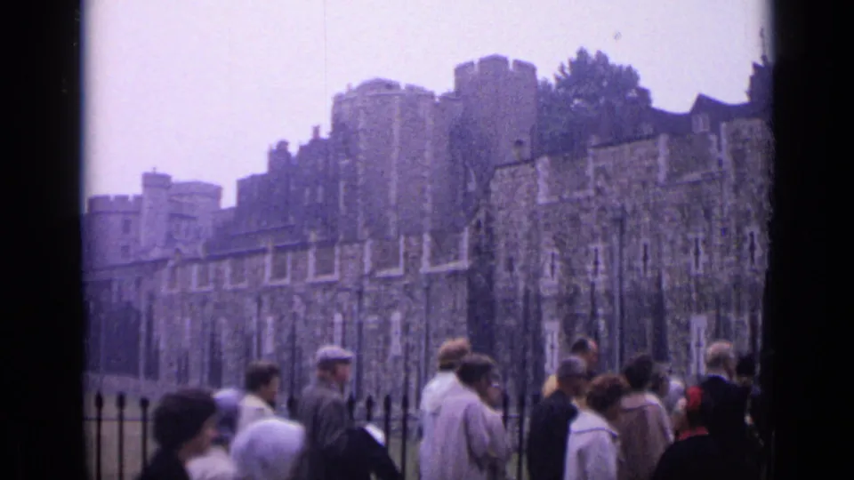 Handheld 8mm view of tourists walking beside the iron fence with the Tower of London's stone exterior in the background.