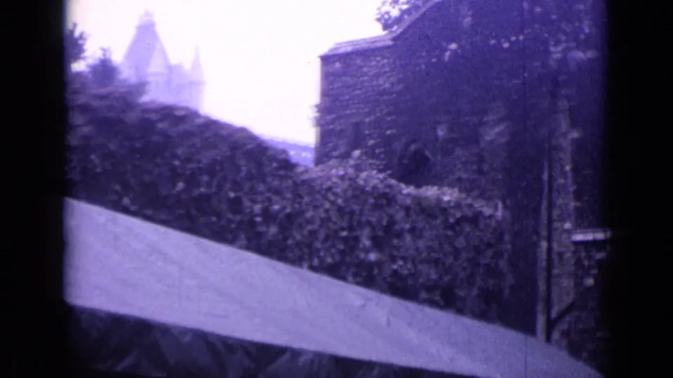 1962 London 8mm frame showing a pale path wall, hedge, stone building edge, and distant tower-like masonry in cloudy light.