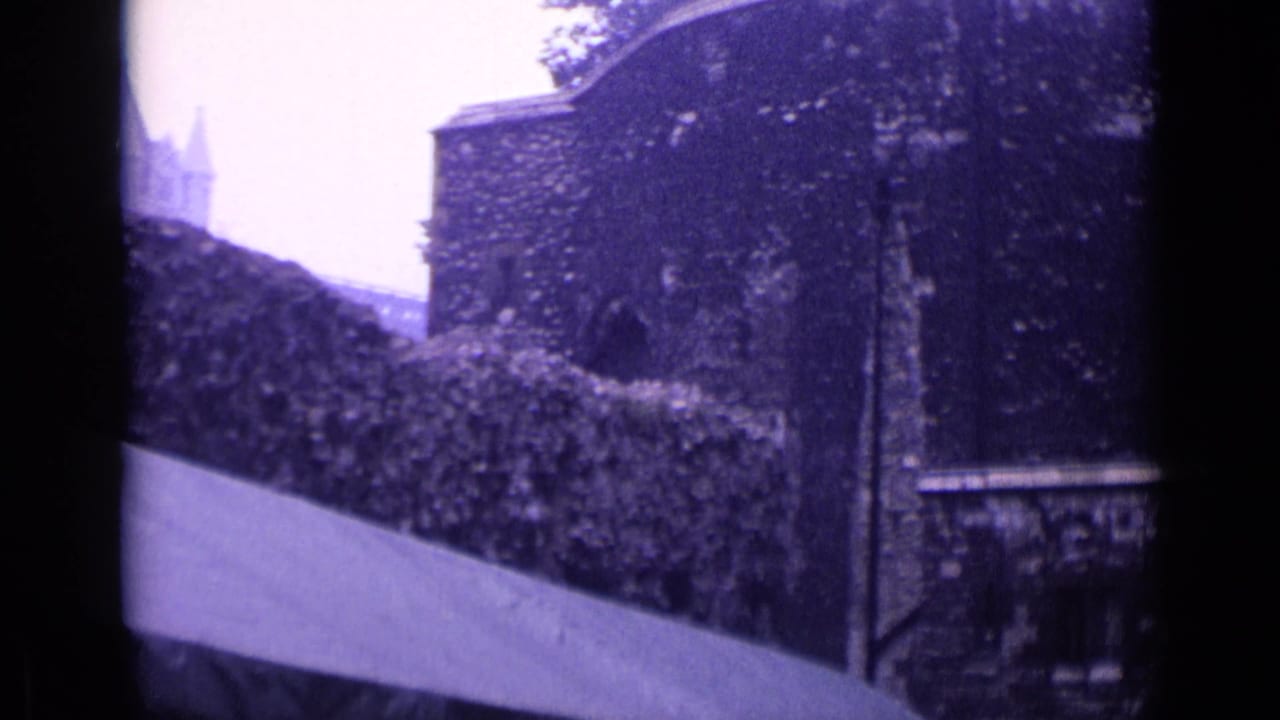 1962 London 8mm frame showing a weathered castle-like exterior wall, hedge line, arched opening, and cloudy sky.