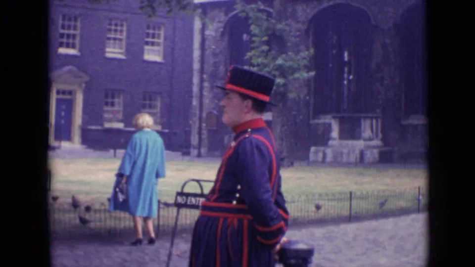 1962 London home-movie frame showing a ceremonial guard by a historic stone building while a woman in a blue coat walks past pigeons.