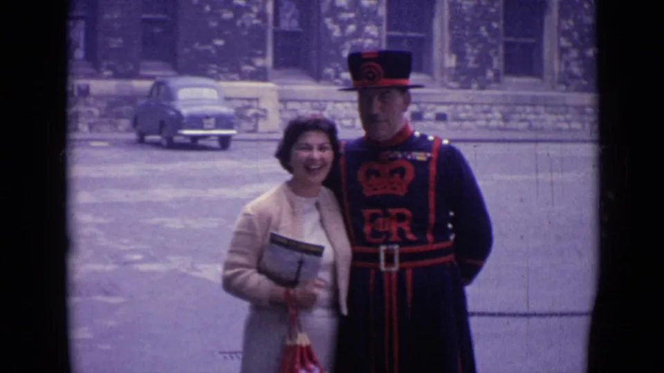 1962 London visitor poses happily with a ceremonial guard in dark uniform, holding a booklet at a historic public site.