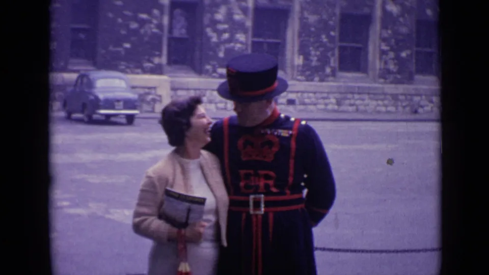London 1962 home-movie frame of a woman laughing or smiling beside a ceremonial guard during a posed tourist photo moment.