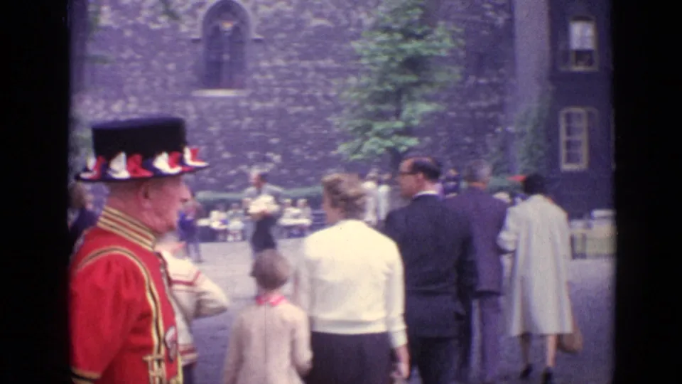 The man in red ceremonial dress moves through a London courtyard as visitors in coats stand and walk nearby.