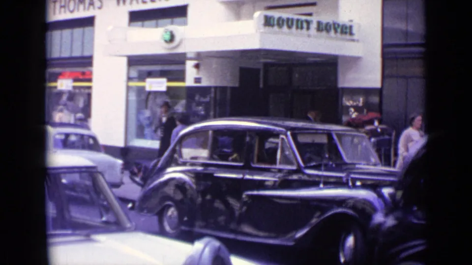 Black vintage limousine in a 1962 London street scene, framed below Mount Royal signage with pedestrians and another car nearby.