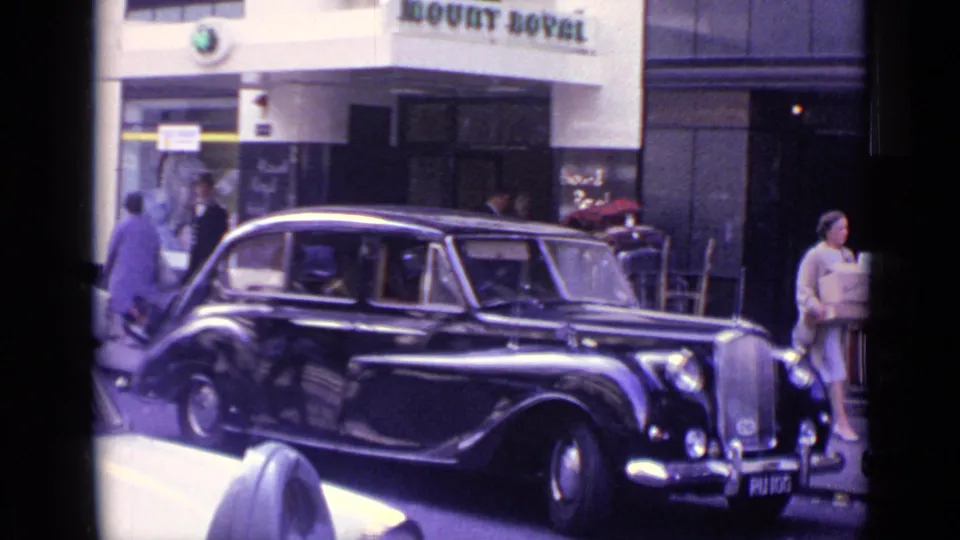 1962 London curbside view with a black vintage limousine, Mount Royal frontage, pedestrians, and a woman carrying a box.