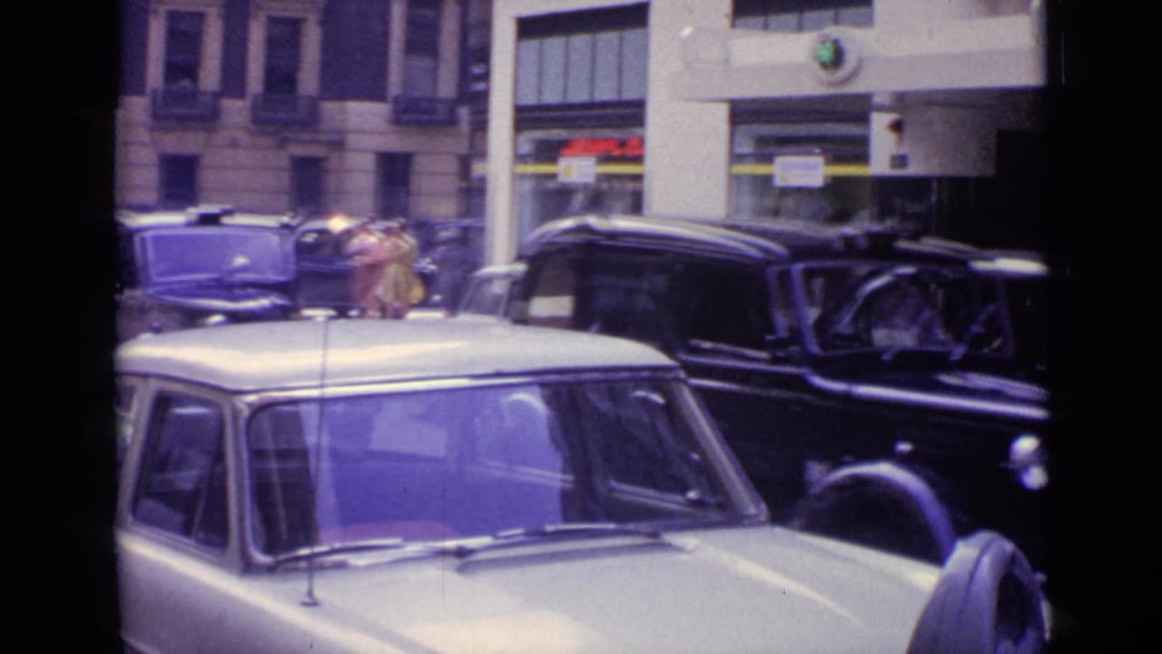 London traffic in 1962 with a pale compact car foreground, black cars, pedestrians, and commercial frontage.