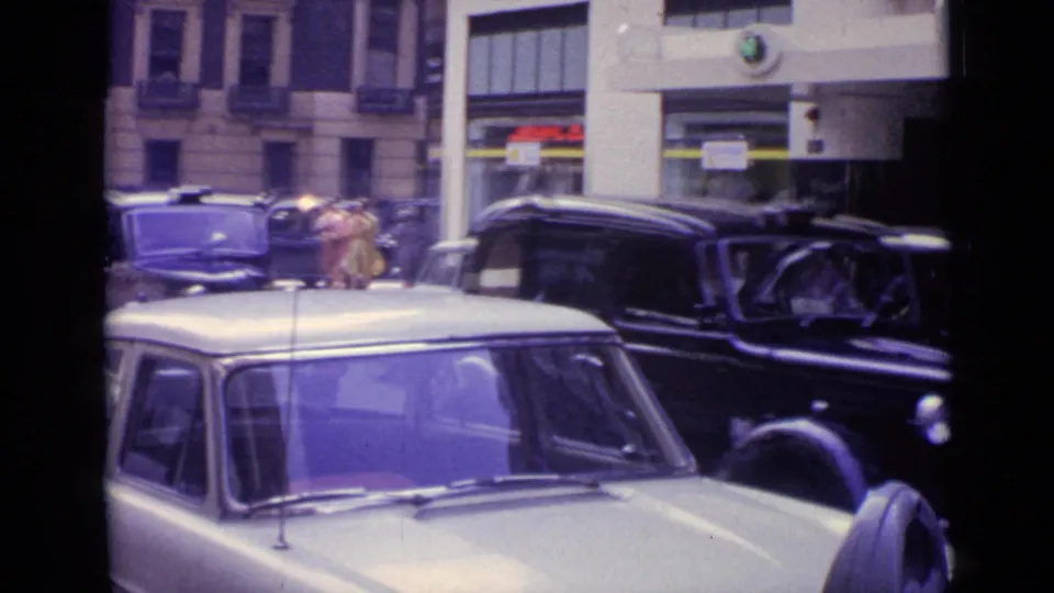 London traffic in 1962 with a pale compact car foreground, black cars, pedestrians, and commercial frontage.