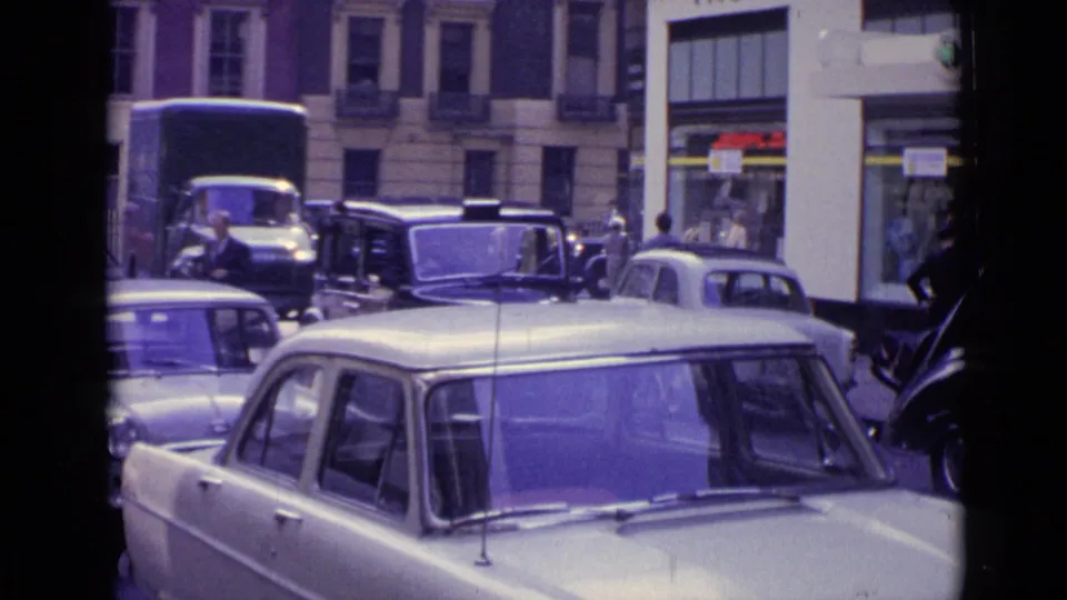 Street-level 1962 London traffic view with a pale compact car, older vehicles, pedestrians, and tall building facades.