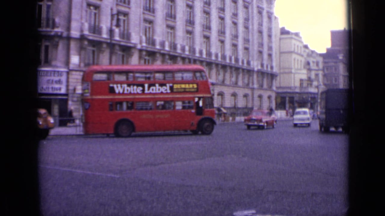 Red double-decker bus crossing a London street with stone buildings and cars behind it.