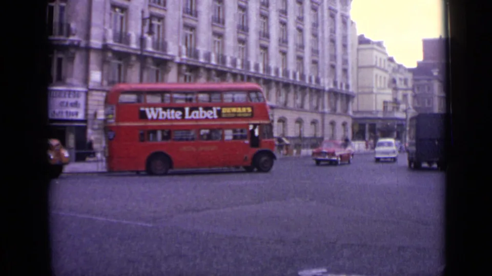 1962 London street view with a red double-decker bus carrying a White Label advertisement, cars, pedestrians, and ornate buildings.