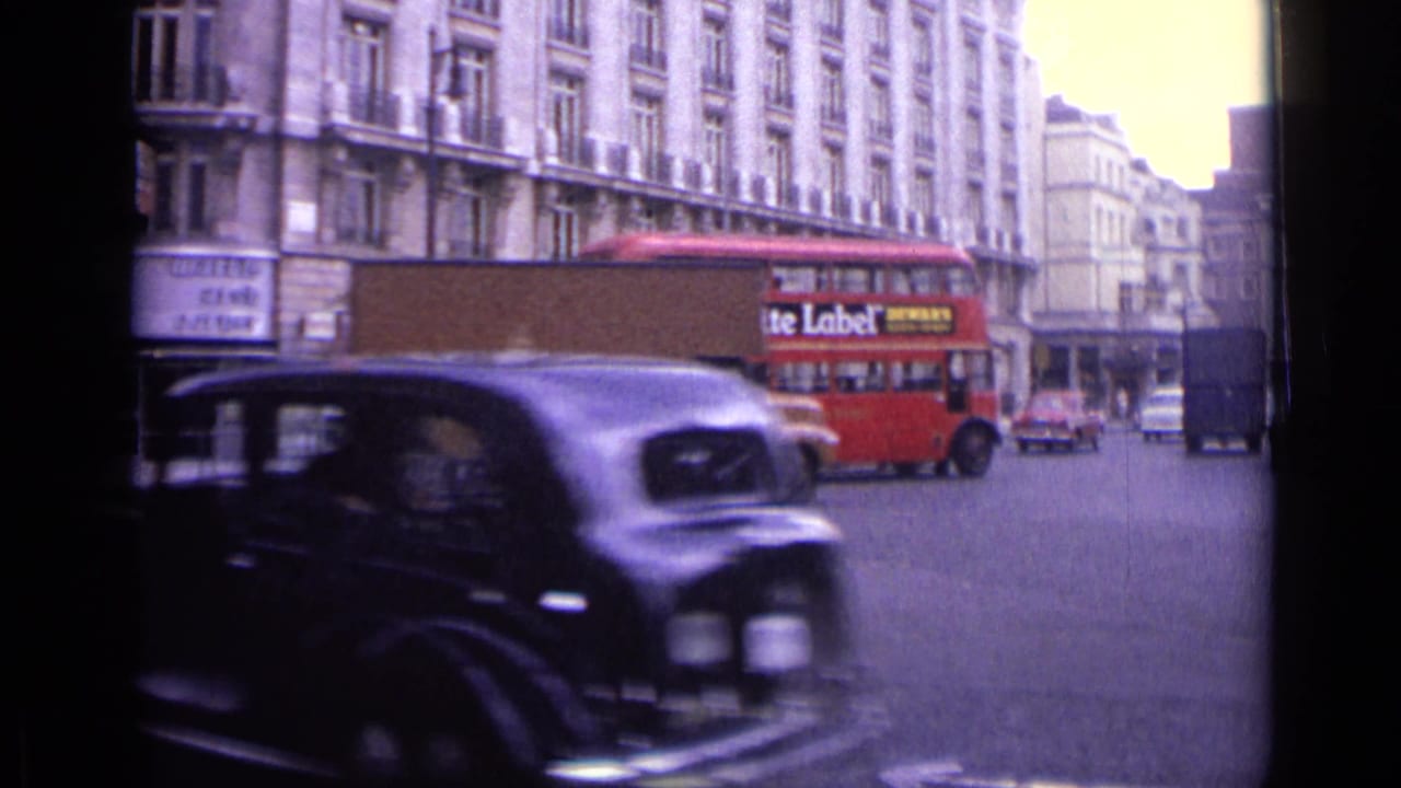 A dark vintage car moves across the foreground as a red London double-decker bus and brown delivery van pass behind it.
