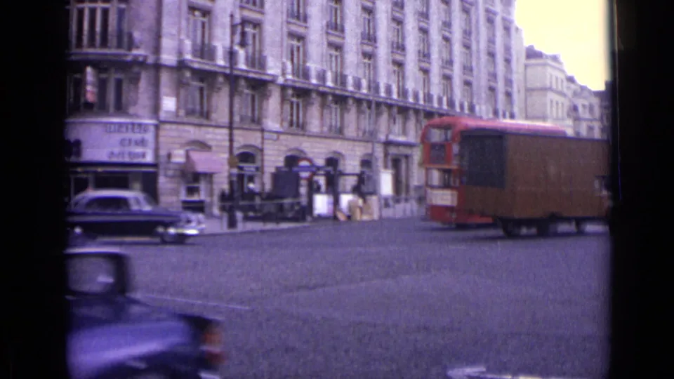 London street corner in 1962 with a red double-decker bus partly hidden by a brown delivery van, shopfronts, cars, and pedestrians.
