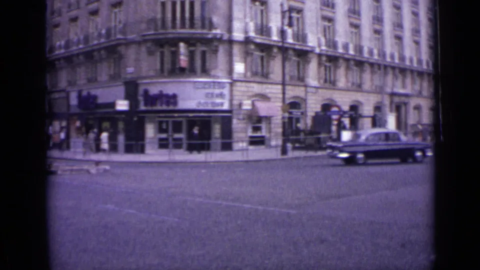 1962 London street corner with pedestrians near shopfronts, a dark car passing, and multi-story stone buildings in view.