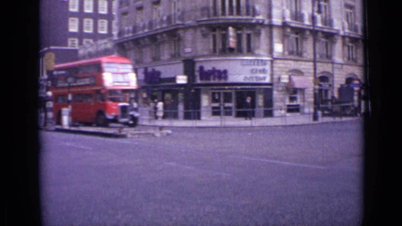 Red double-decker bus passing a London street corner with shopfronts and pedestrians.