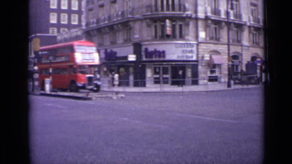 1962 London street view with a red double-decker bus, older buildings, shopfronts, pedestrians, and traffic island details.