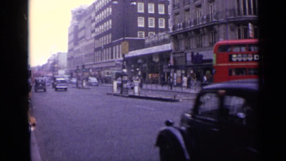 1962 London street traffic with older cars, pedestrians near shops, and a red double-decker bus partly visible at the edge.