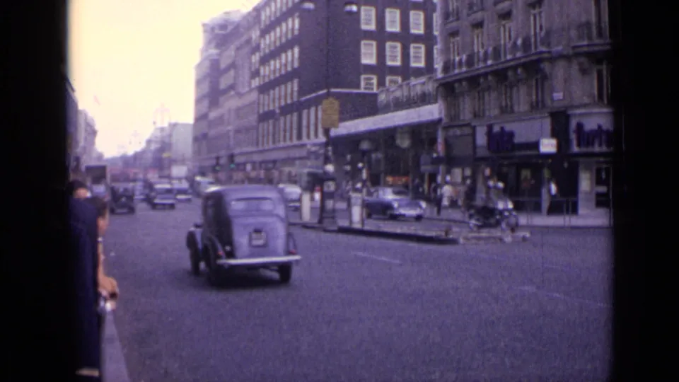 1962 London street view with a dark older car, pedestrians, shopfronts, bollards, and multi-story buildings in soft 8mm color.