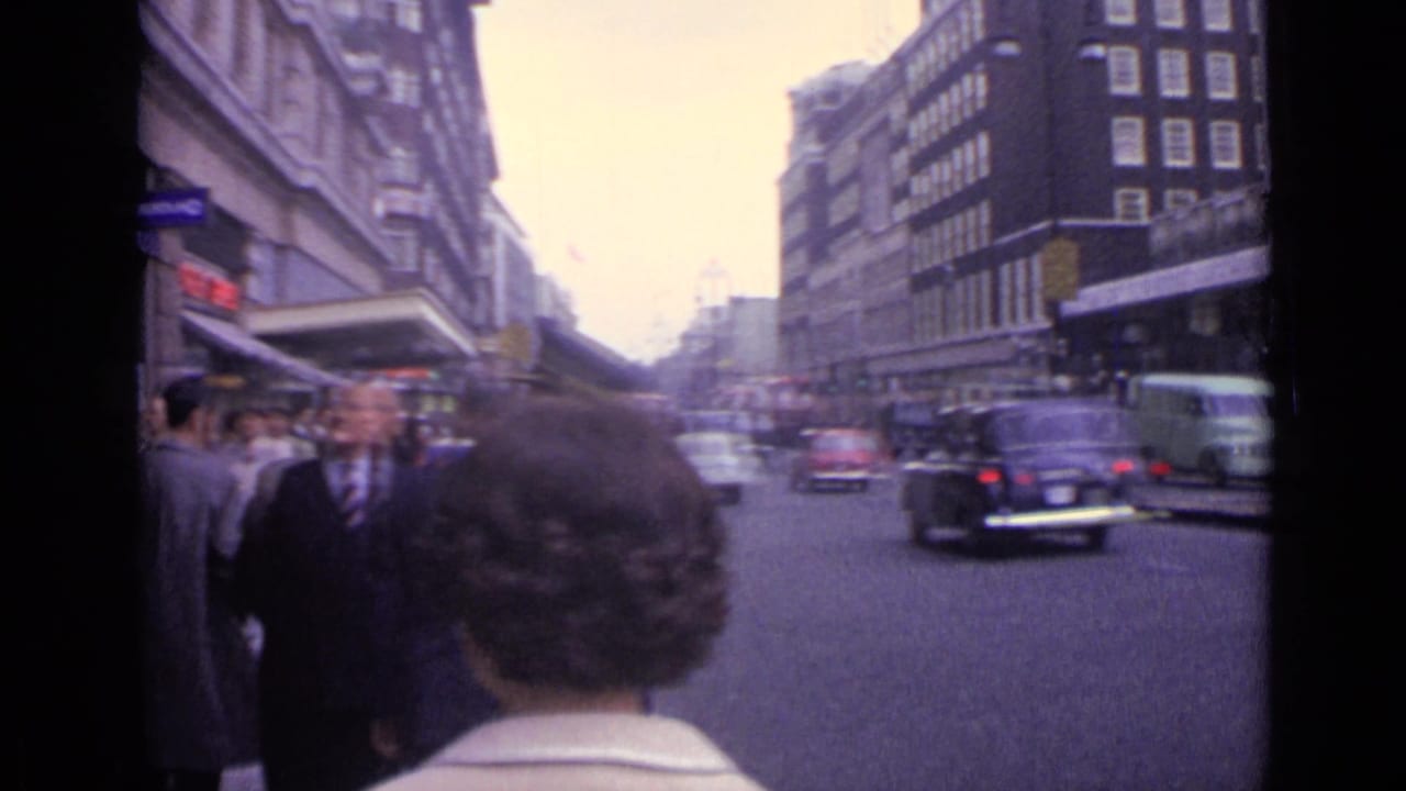 Pedestrians wait at a curb as cars and a van pass on a London street.