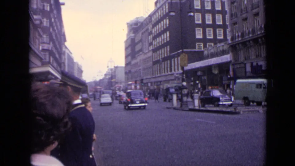 1962 London traffic scene with pedestrians at the edge of frame, cars and a van on the road, and commercial buildings beyond.