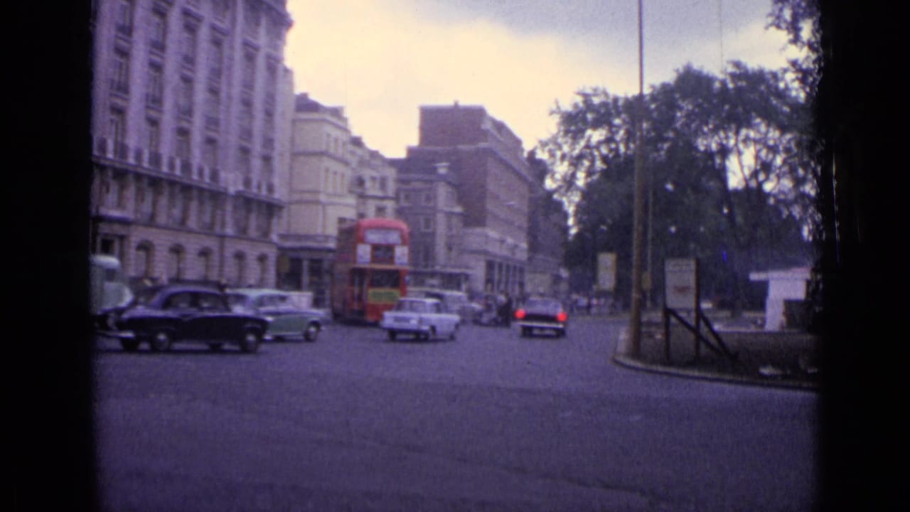 1962 London street with a red double-decker bus, mid-century cars, tall buildings, trees, and roadside signs.