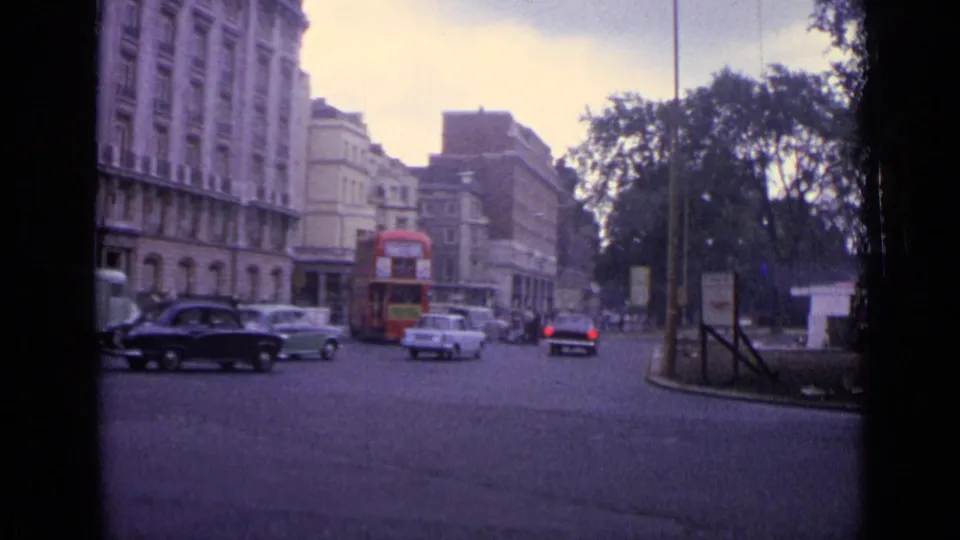 1962 London street with a red double-decker bus, mid-century cars, tall buildings, trees, and roadside signs.