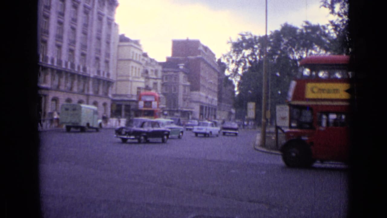 1962 London junction with a red double-decker bus at right, cars and vans in traffic, buildings, trees, and street signs.
