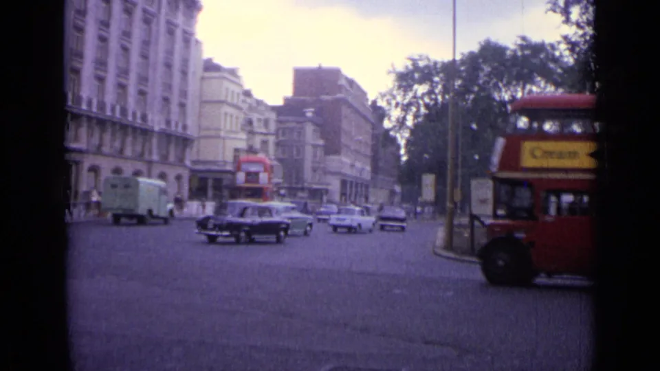 1962 London junction with a red double-decker bus at right, cars and vans in traffic, buildings, trees, and street signs.