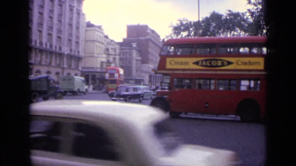 1962 London street view with a red double-decker bus, vintage cars, and period buildings seen from a handheld 8mm camera.
