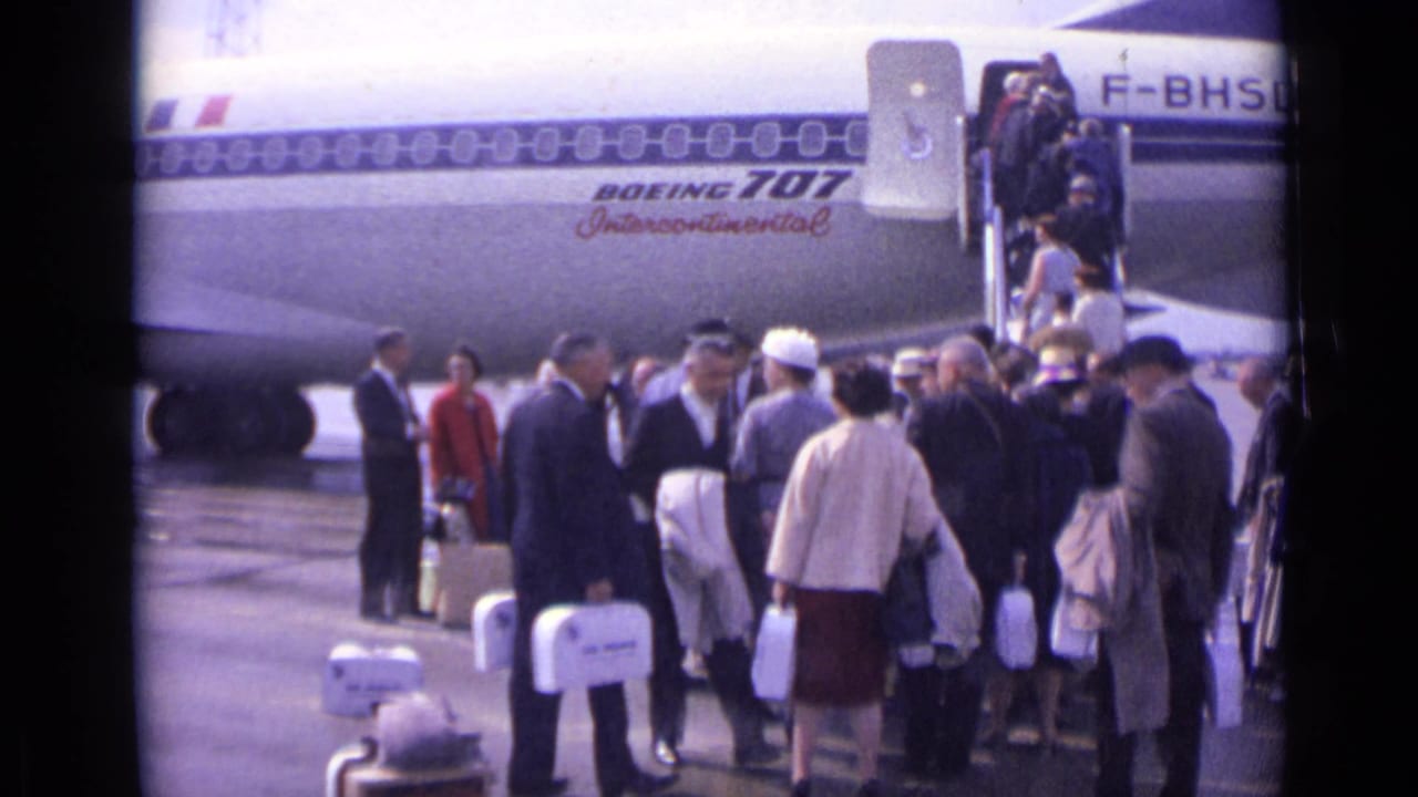 Passengers with luggage gather beside a Boeing 707 as others board by stairs.