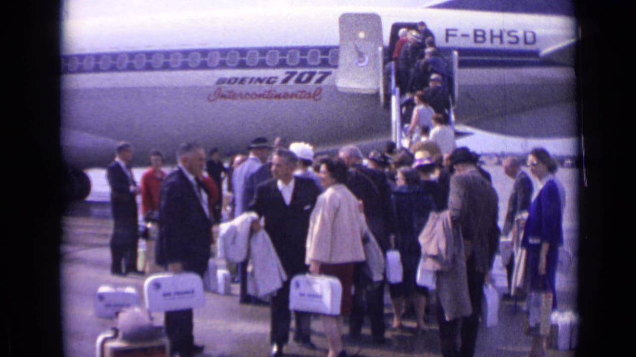 Passengers in 1960s dress wait and converse near Air France-marked luggage while others climb the Boeing 707 stairs on a London airport tarmac.