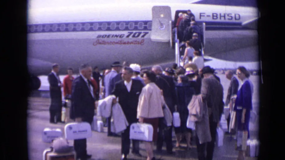 Passengers in 1960s dress wait and converse near Air France-marked luggage while others climb the Boeing 707 stairs on a London airport tarmac.