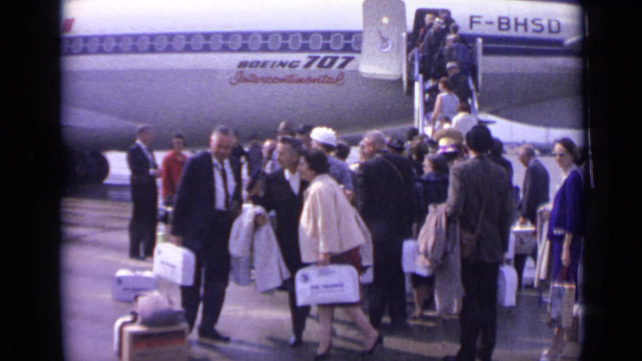 A man in a suit moves through the foreground as passengers cluster with bags beneath the Boeing 707 Intercontinental marking in the 1962 London airport scene.