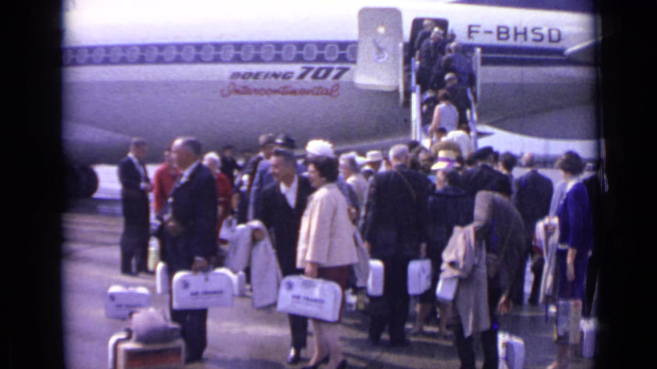 Passengers queue on the tarmac beside a Boeing 707 with boarding stairs and white travel cases.