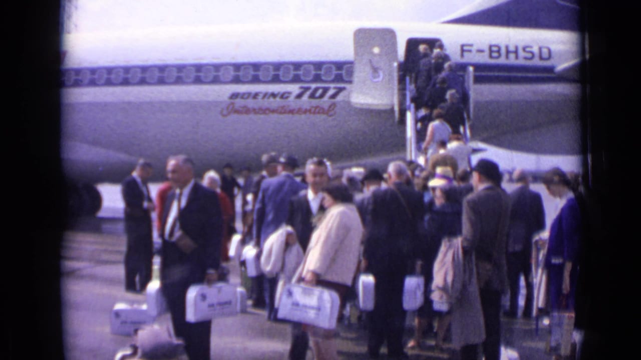 Passengers in 1960s clothing wait and move toward the stairs of a Boeing 707 at a London airport tarmac.