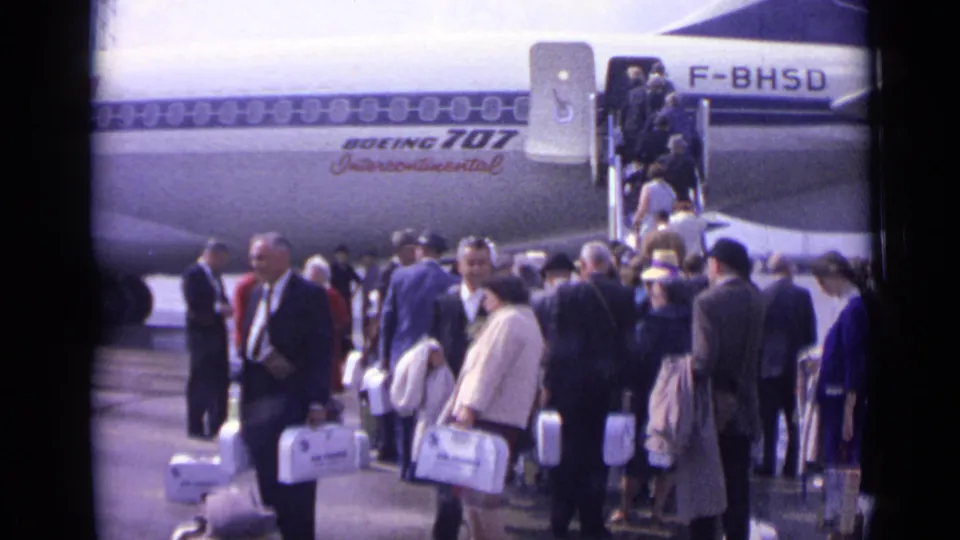 Passengers in 1960s clothing wait and move toward the stairs of a Boeing 707 at a London airport tarmac.