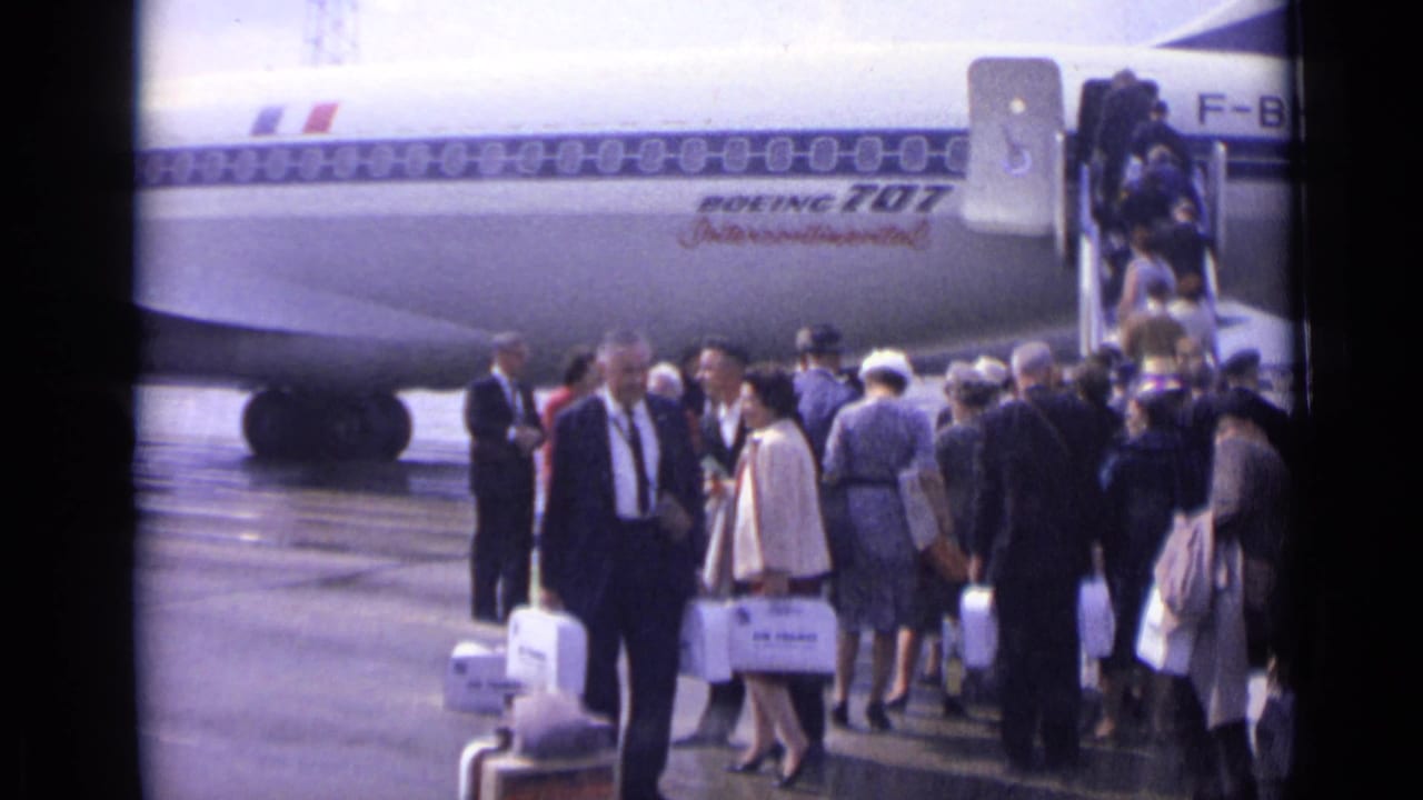 Passengers gather beside a Boeing 707 with boarding stairs on an airport apron.