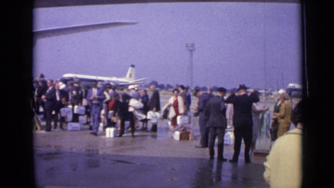 Passengers at London Airport stand with cases on the open apron, with aircraft and airport towers in the background.