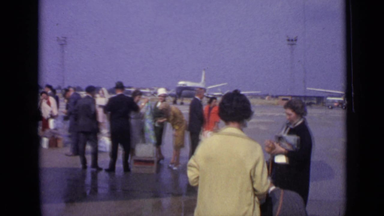 1960s travelers in coats and suits cross the London airport apron with luggage near parked aircraft and service vehicles.