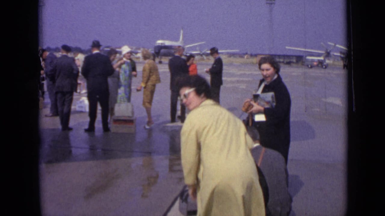 A woman in a yellow coat handles luggage on the London Airport apron as other passengers wait near distant aircraft.