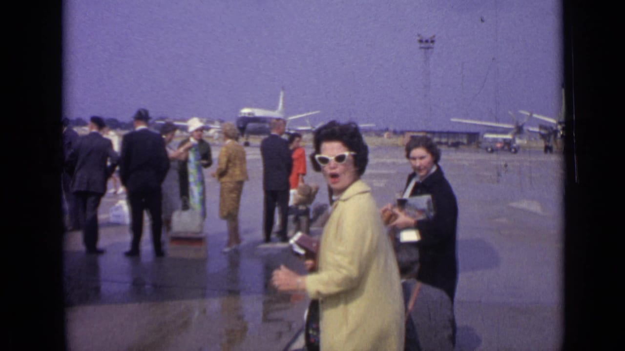 Passengers with luggage stand on an airport tarmac near parked airliners.