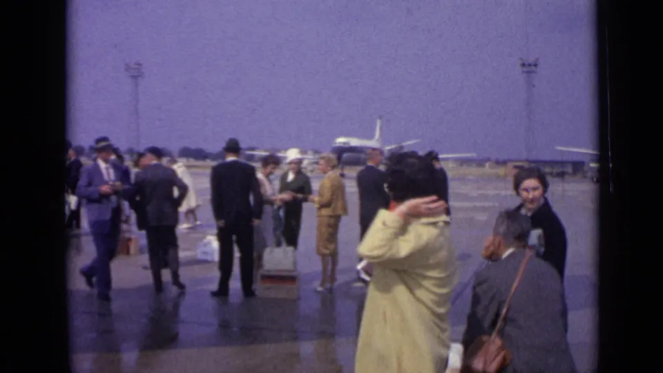1962 London airport tarmac scene with travelers in coats and suits, luggage on the ground, and vintage airliners behind them.