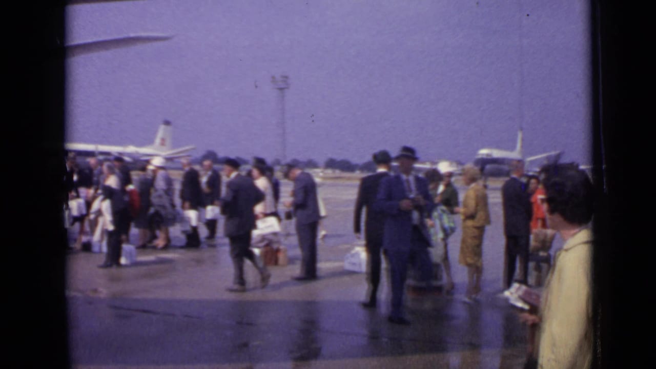 1962 London frame showing suited passengers, luggage, wet pavement, and a propeller aircraft in the background on the airport tarmac.