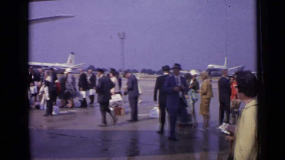 1962 London frame showing suited passengers, luggage, wet pavement, and a propeller aircraft in the background on the airport tarmac.