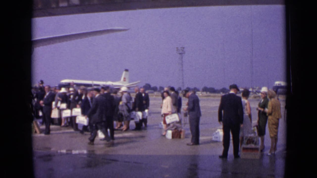 1962 London home-movie frame with passengers spread across a wet airport tarmac, luggage at their feet, and airliners beyond.