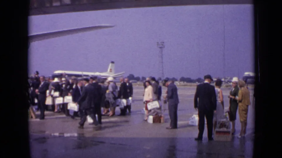 1962 London home-movie frame with passengers spread across a wet airport tarmac, luggage at their feet, and airliners beyond.