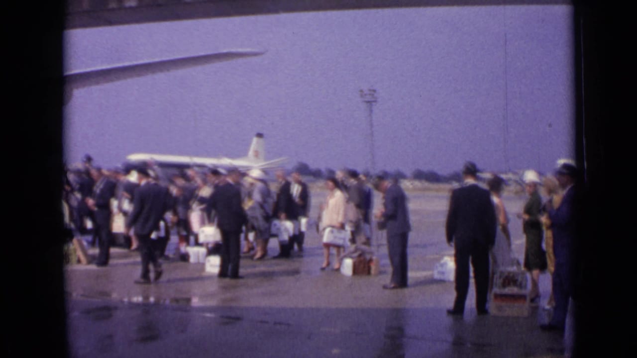 Passengers with luggage wait on a wet airport tarmac near a jet airliner.