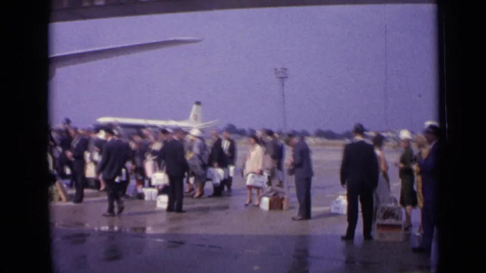 1962 London airport scene with suited passengers and luggage waiting on wet tarmac near a mid-century jet airliner.