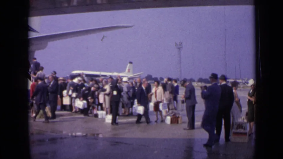Passengers in 1960s dress cluster with suitcases on a London airport tarmac as a jet airliner waits behind them.