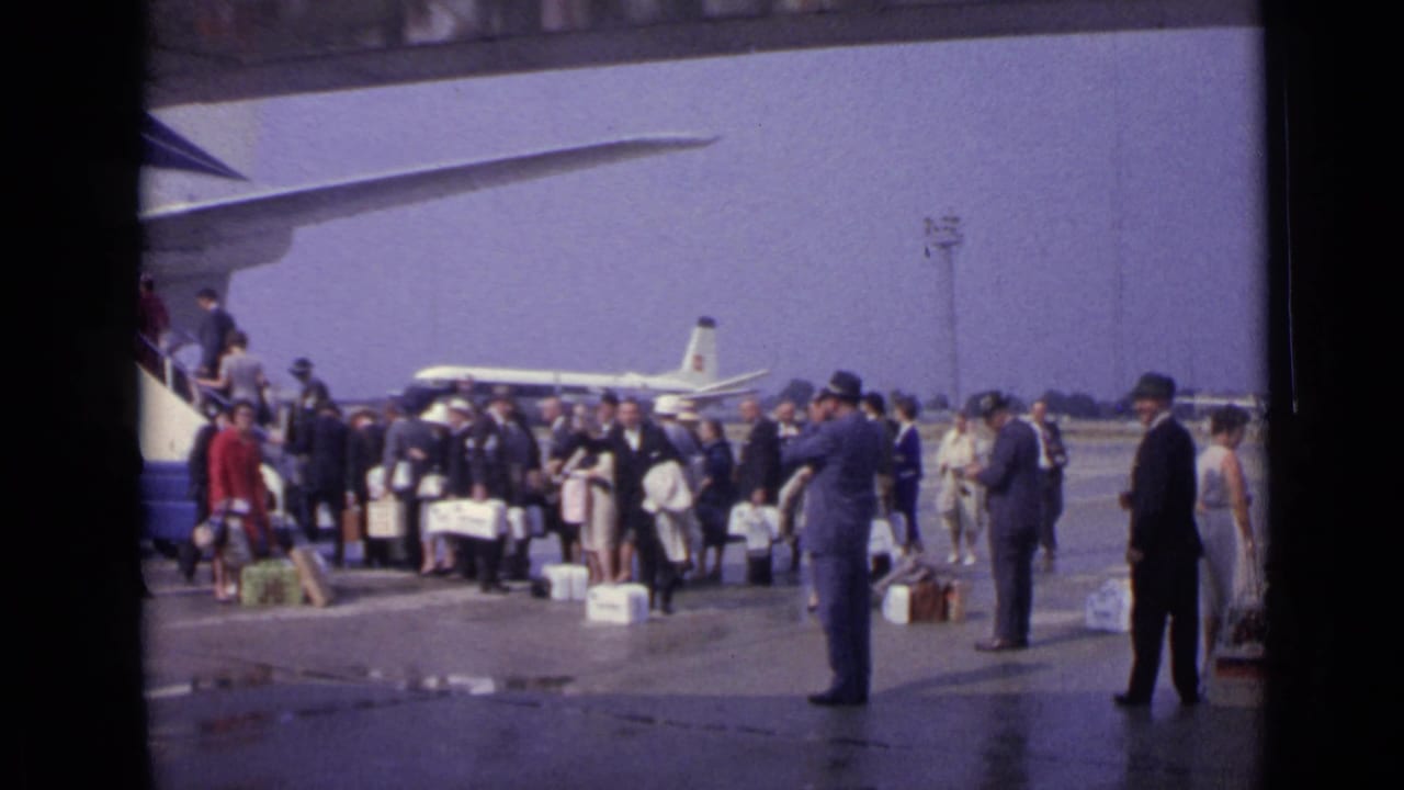 Travelers stand with luggage near aircraft stairs while another plane sits in the background.
