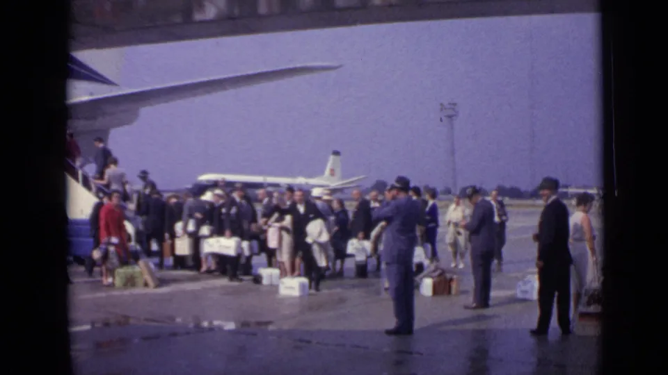 1962 London airport travelers wait with suitcases near Boeing 707 boarding stairs, with another aircraft visible beyond them.